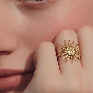 May include: A gold-colored ring featuring a sun design with a face and radiating rays. The ring is worn on a finger, with the sun's face adorned with small, sparkling details. The ring is a piece of jewelry.