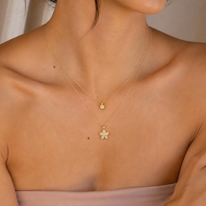 May include: A layered gold necklace set featuring two delicate chains. The shorter chain holds a small, round, clear stone pendant, while the longer chain features a five-pointed star-shaped pendant encrusted with small, clear, sparkling stones.