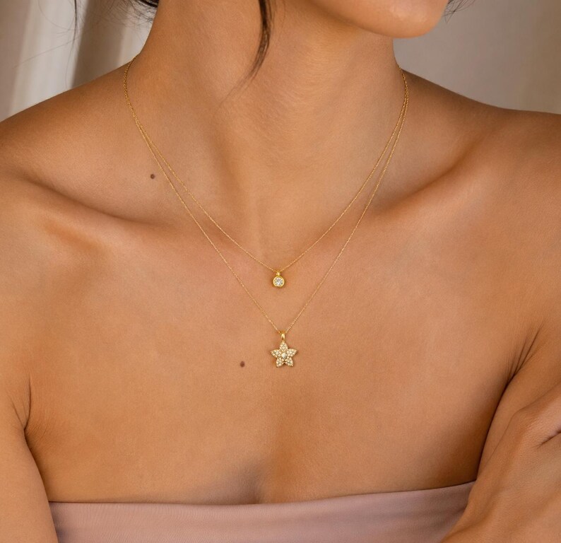 May include: A layered gold necklace set featuring two delicate chains. The shorter chain holds a small, round, clear stone pendant, while the longer chain features a five-pointed star-shaped pendant encrusted with small, clear, sparkling stones.