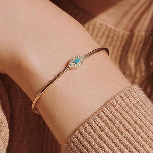 Könnte beinhalten: Ein goldfarbenes Armband mit einem zentralen, augenförmigen Anhänger. Der Anhänger ist mit kleinen, klaren Steinen besetzt, die einen einzelnen, kleinen, runden, leuchtend blauen Stein in der Mitte umgeben und so einen Blickfang bilden.
