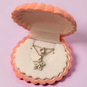 May include: A gold necklace featuring a star-shaped pendant encrusted with small clear stones and a single round bezel-set stone on the chain, presented inside an open peach-colored, shell-shaped jewelry box with a white velvet interior lining.