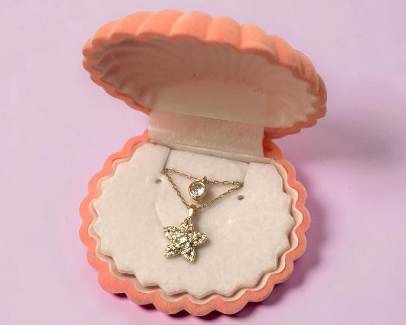 May include: A gold necklace featuring a star-shaped pendant encrusted with small clear stones and a single round bezel-set stone on the chain, presented inside an open peach-colored, shell-shaped jewelry box with a white velvet interior lining.