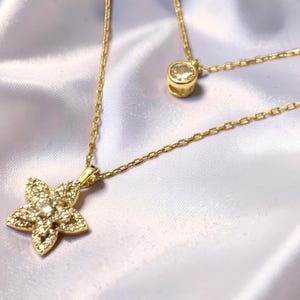 May include: A gold-toned layered necklace featuring two delicate chains. The shorter chain holds a single round clear stone pendant, while the longer chain features a five-petaled flower pendant encrusted with small, sparkling clear stones.