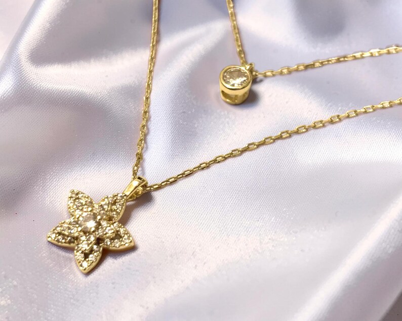 May include: A gold-toned layered necklace featuring two delicate chains. The shorter chain holds a single round clear stone pendant, while the longer chain features a five-petaled flower pendant encrusted with small, sparkling clear stones.