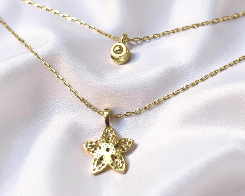 May include: A gold-toned layered necklace featuring two delicate chains. The upper chain holds a small circular charm, while the lower chain features a five-pointed star pendant with a textured, perforated surface and a central clear stone.