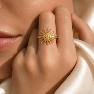 May include: A gold-toned ring featuring a central sun motif with a detailed face and radiating rays. The band of the ring has a textured, hammered finish, and the sun design is positioned prominently on the front of the circular metal band.