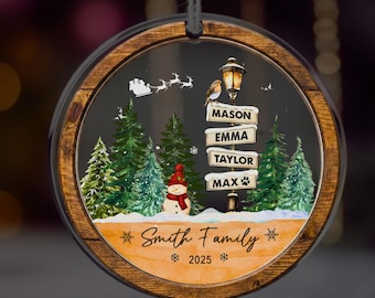 May include: A round Christmas ornament with a wooden frame. Inside, a winter scene features a snowman, pine trees, and a signpost with names. The ornament reads "Smith Family 2025". A black string is attached for hanging.