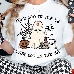 May include: White t-shirt with a Halloween-themed graphic. The design features a ghost wearing a nurse's hat, a pumpkin, a spider web, and the text "CODE BOO IN THE ER". The shirt is paired with black and white patterned pants.