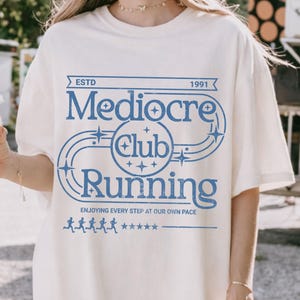 May include: Cream-colored t-shirt with a blue graphic that reads "Mediocre Club Running" above the words "Enjoying every step at our own pace." The shirt also features a design of running figures and stars.