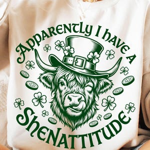 Op de afbeelding: Witte sweatshirt met een groene afbeelding van een Schotse Hooglander die een hoge hoed draagt ​​versierd met klavers. De tekst "Apparently I have a Shenattitude" is rond de koe gebogen. Klaver- en muntgrafieken zijn verspreid.