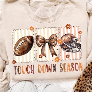 Puede incluir: Sudadera beige con un diseño de fútbol americano, lazo y casco. El fútbol, el lazo y el casco están en marcos rectangulares separados con un fondo a rayas. El texto "TOUCH DOWN SEASON" está impreso debajo en marrón y naranja.