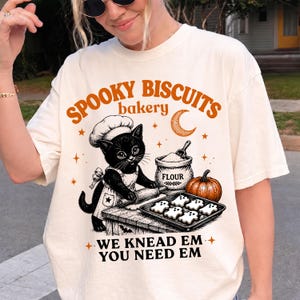 May include: A cream-coloured t-shirt featuring a graphic of a black cat wearing a chef hat and apron, rolling dough. The text reads SPOOKY BISCUITS bakery WE KNEAD EM YOU NEED EM, with illustrations of a flour bag, a pumpkin, and ghost-shaped cookies.