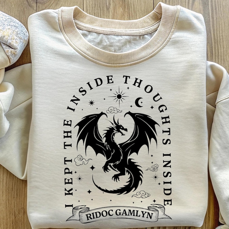 May include: Cream-colored sweatshirt with a black dragon graphic. The design includes the text "I KEPT THE INSIDE THOUGHTS INSIDE" and "RIDOC GAMLYN" on a banner. The dragon is surrounded by stars, clouds, a sun, and a crescent moon.