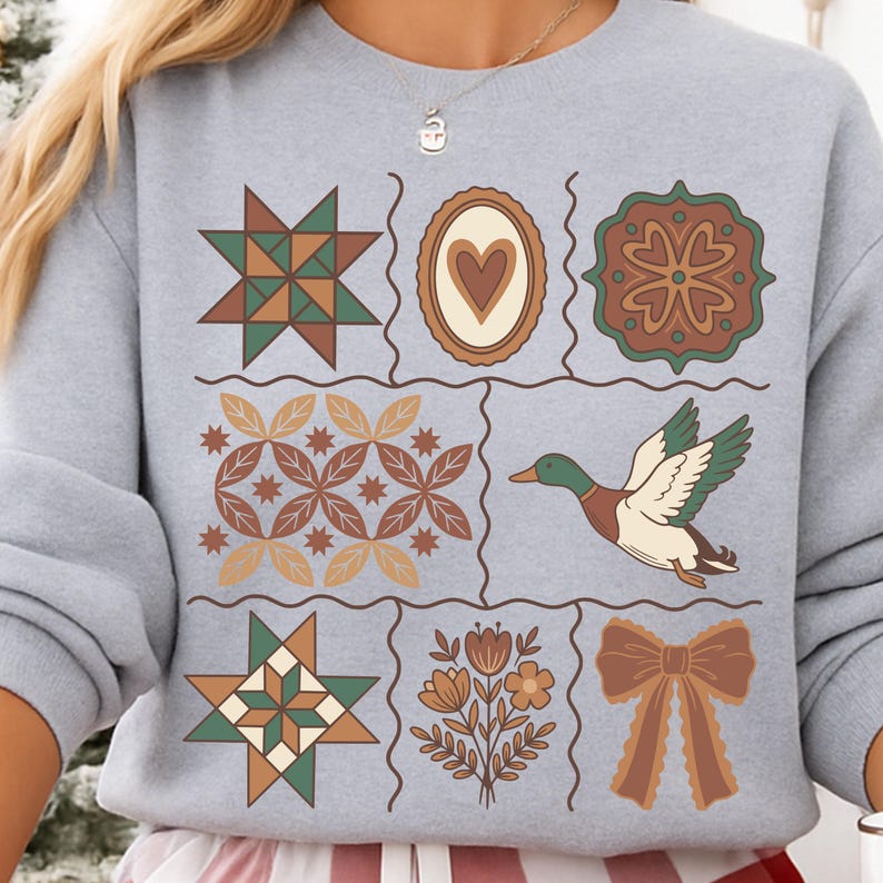 May include: A pale grey sweatshirt with a patchwork design featuring geometric and floral patterns in brown, green, and tan. A flying duck graphic is also included. The design is reminiscent of a quilt.