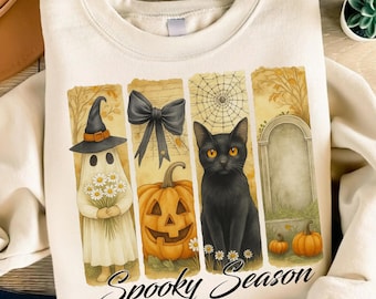 May include: Cream-colored sweatshirt featuring a Halloween-themed graphic. The design includes a ghost, jack-o'-lantern, black cat, and tombstone, with the words "Spooky Season" in a script font. The artwork has a watercolor style.