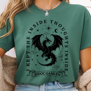 May include: Green t-shirt with a black dragon graphic. The design includes the text "I KEPT THE INSIDE THOUGHTS INSIDE" and "RIDOC GAMLYN" on a banner. The dragon has large wings and is surrounded by stars, clouds, a sun, and a crescent moon.