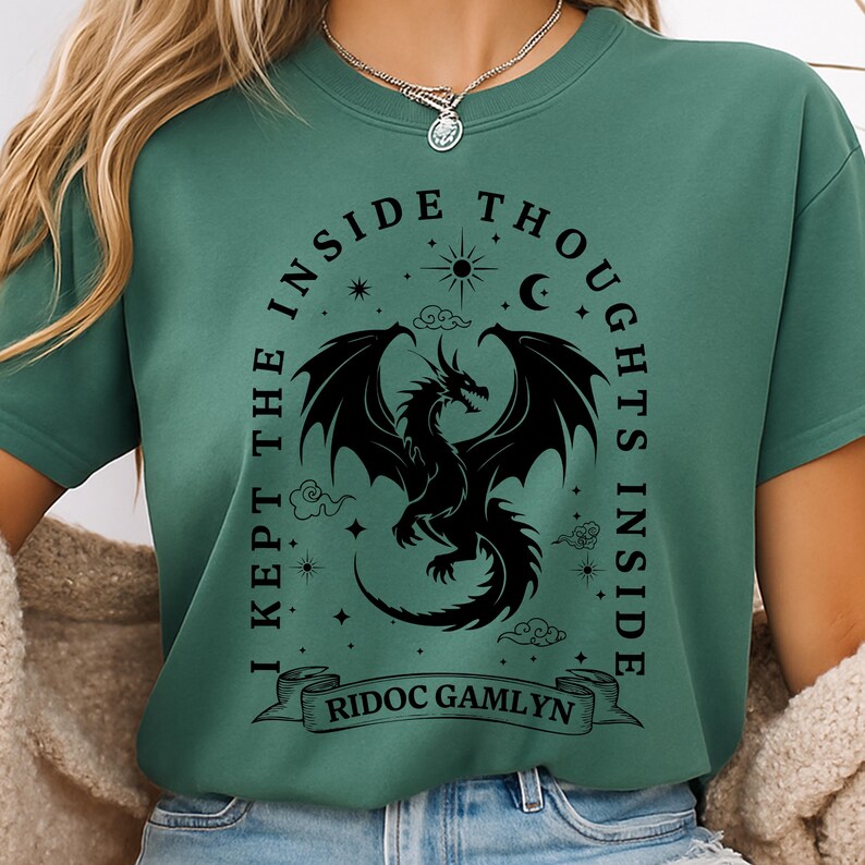 May include: Green t-shirt with a black dragon graphic. The design includes the text "I KEPT THE INSIDE THOUGHTS INSIDE" and "RIDOC GAMLYN" on a banner. The dragon has large wings and is surrounded by stars, clouds, a sun, and a crescent moon.