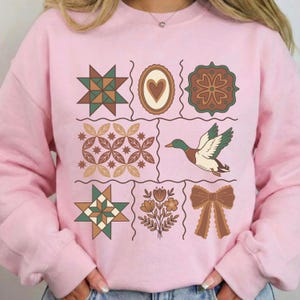 May include: A pale pink sweatshirt with a grid of nine decorative elements. The elements include a duck, a heart, a bow, and various geometric and floral designs in green, brown, and beige. The sweatshirt is a casual style.