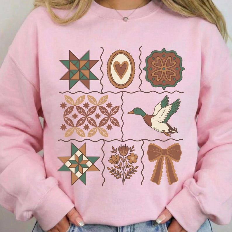 May include: A pale pink sweatshirt with a grid of nine decorative elements. The elements include a duck, a heart, a bow, and various geometric and floral designs in green, brown, and beige. The sweatshirt is a casual style.