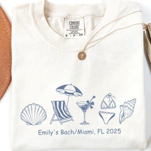 Può includere: T-shirt color crema con un design a tema spiaggia in blu. Il design include una conchiglia, una sedia a sdraio, un ombrellone, un cocktail, un bikini e una conchiglia. Il testo "Emily's Bach/Miami, FL 2025" è stampato sotto. Una collana d'oro è drappeggiata sulla maglietta.