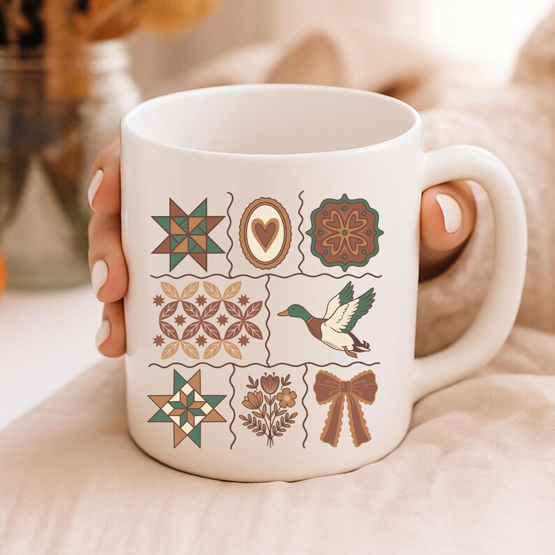 May include: White ceramic mug with a patchwork design featuring stars, hearts, and a duck in flight. The mug has a large handle and is held by a person.