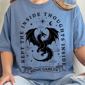 May include: Light blue t-shirt featuring a black dragon design with the text "I KEPT THE INSIDE THOUGHTS INSIDE" and "RIDOC GAMLYN". The design includes stars, a crescent moon, and clouds.
