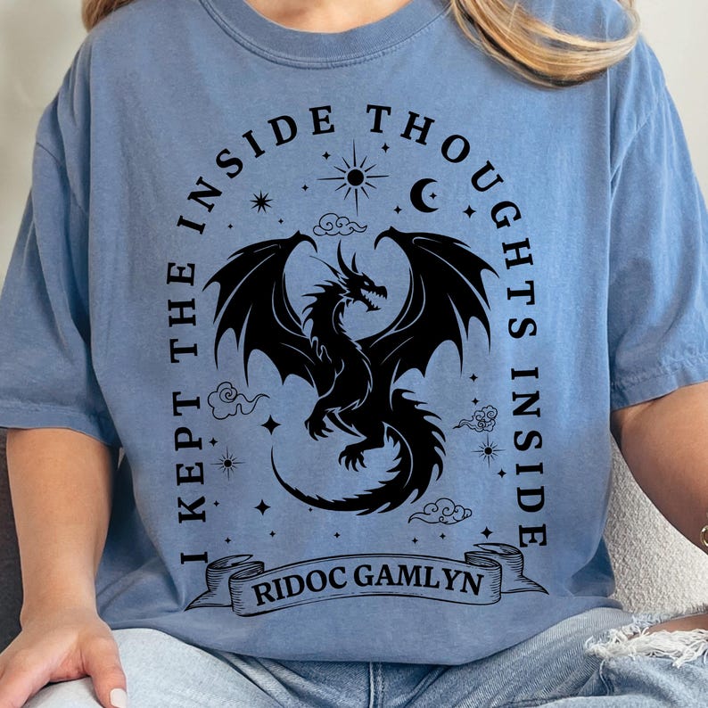 May include: Light blue t-shirt featuring a black dragon design with the text "I KEPT THE INSIDE THOUGHTS INSIDE" and "RIDOC GAMLYN". The design includes stars, a crescent moon, and clouds.