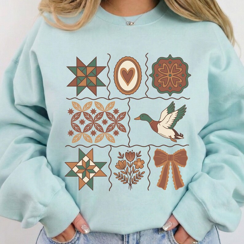 May include: A pale blue sweatshirt with a patchwork design. The design features a star, a heart, a floral pattern, a duck, and a bow, in shades of brown, green, and cream. The overall style is vintage.