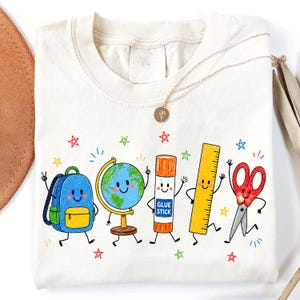 May include: A cream-colored t-shirt featuring a colorful graphic of five anthropomorphic school supplies: a blue backpack, a globe, an orange glue stick labeled GLUE STICK, a yellow ruler, and red scissors, all surrounded by small multi-colored stars.