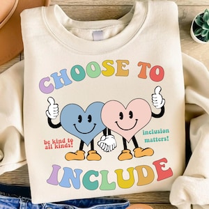 May include: Cream-colored sweatshirt with the words "CHOOSE TO INCLUDE" in colorful, retro-style lettering. Two smiling heart characters hold hands, with thumbs up. Text reads "be kind to all kinds!" and "inclusion matters!"