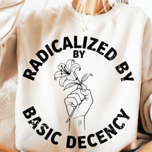 May include: Cream-colored sweatshirt with the text "RADICALIZED BY BASIC DECENCY" in a circular design. A black and white illustration of a hand holding a lily is in the center of the text. The sweatshirt is a casual style.