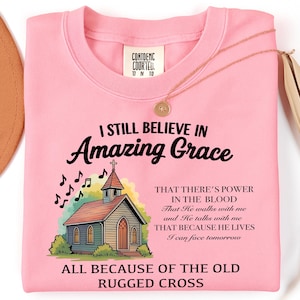 Può includere: Una felpa rosa chiaro con la scritta "I Still Believe in Amazing Grace" e un disegno di una chiesa. Una collana d'oro con un ciondolo rotondo è drappeggiata sulla maglia.