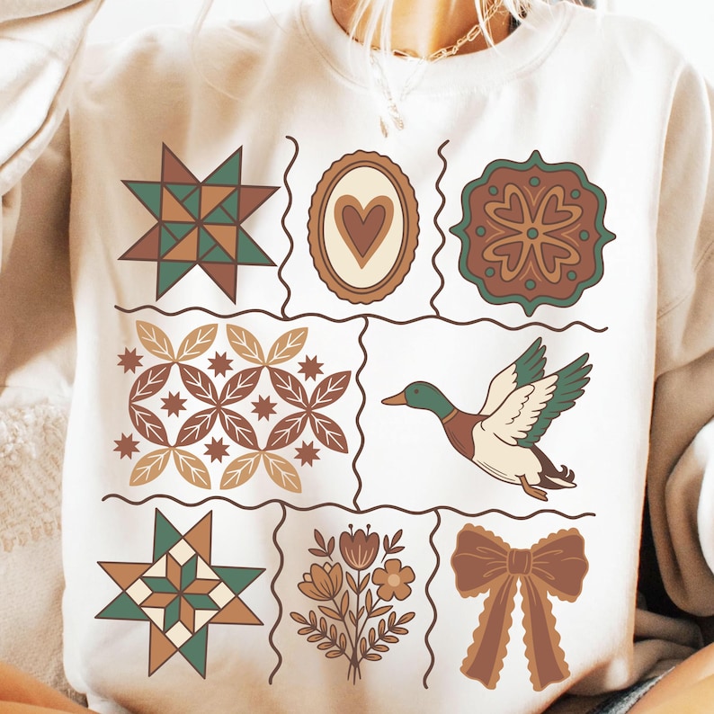 May include: Cream-coloured sweatshirt with a grid of nine decorative squares. The squares display various designs, including a duck, floral arrangements, geometric patterns, and a bow. The colour palette includes shades of brown, green, and cream.