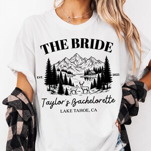 May include: White t-shirt with a black graphic design featuring the text "THE BRIDE" above a mountain and lake scene with deer and trees. The text "Taylor's Bachelorette" and "LAKE TAHOE, CA" are also included.