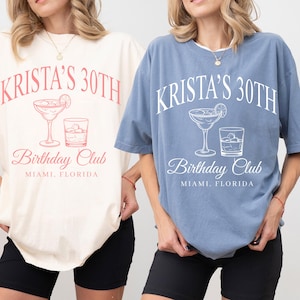 Op de afbeelding: Twee oversized T-shirts, één crème en één blauw, met de tekst "KRISTA'S 30TH" boven een cocktailglas en een whiskyglas. Daaronder staat de tekst "Birthday Club" en "MIAMI, FLORIDA". De shirts worden gedragen met zwarte shorts.