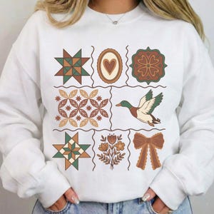 May include: White sweatshirt with a patchwork design featuring geometric and nature-inspired illustrations. The design includes stars, hearts, flowers, and a duck in shades of green, brown, and beige. The sweatshirt has long sleeves and a crew neck.