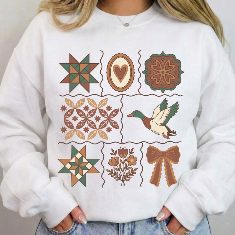 May include: White sweatshirt with a patchwork design featuring geometric and nature-inspired illustrations. The design includes stars, hearts, flowers, and a duck in shades of green, brown, and beige. The sweatshirt has long sleeves and a crew neck.