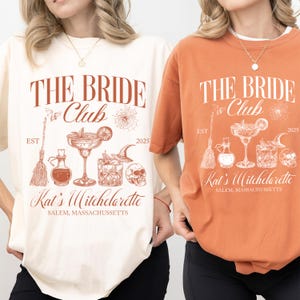 Könnte beinhalten: Zwei T-Shirts, eines in Creme und eines in Orange, mit dem Text "THE BRIDE Club" und einer Hexen-Grafik. Das Design umfasst einen Besen, eine Zaubertrankflasche, einen Cocktail, einen Hexenhut und einen Totenkopf. Der Text "Kat's Witchelarette, Salem, Massachusetts" ist ebenfalls aufgedruckt.