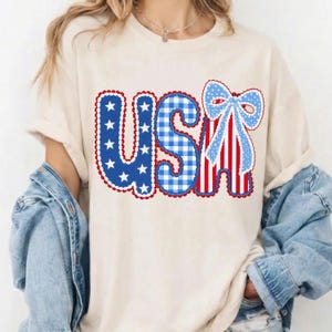 May include: Cream-colored t-shirt featuring the word "USA" in a patriotic design. The letters are decorated with blue stars, gingham, and red and white stripes, with a blue bow accent. The shirt is worn with a denim jacket.