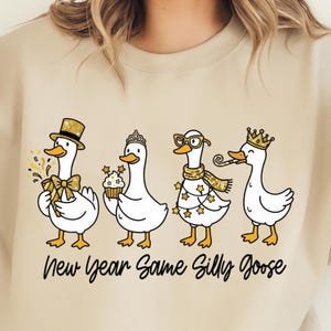 May include: Beige sweatshirt with a graphic of four cartoon geese, each adorned with festive accessories like a top hat, tiara, glasses, and crown. The text "New Year Same Silly Goose" is printed below the geese in black script.