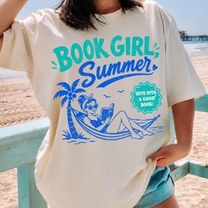 Op de afbeelding: Crèmekleurig T-shirt met een blauwe en turquoise afbeelding. De afbeelding toont "BOOK GIRL Summer" met een palmboom en een vrouw in een hangmat die een boek leest. De tekst "DIVE INTO A GOOD BOOK!" is ook opgenomen.