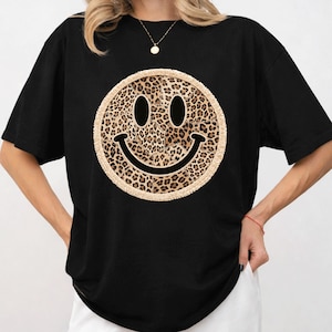 May include: Black t-shirt featuring a large smiley face design. The smiley face has a leopard print pattern and a tan border. The shirt has short sleeves and a crew neck.