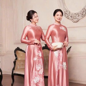 May include: Two women in matching pink satin dresses with long sleeves and floral embroidery. The dresses have high necklines and are floor-length. The dresses are from Amy Bridal.