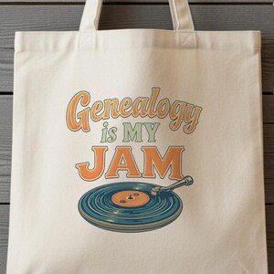 May include: Cream-colored tote bag with the text "Genealogy is my JAM" in orange and brown. Below the text is a teal and orange vinyl record illustration. The bag is hanging on a metal hook against a gray wooden wall.