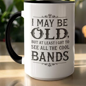 Może przedstawiać: Biały ceramiczny kubek z czarnym uchwytem i obramowaniem. Kubek zawiera napis "I MAY BE OLD, BUT AT LEAST I GOT TO SEE ALL THE COOL BANDS" w czarnych literach. Kubek stoi na jasnej powierzchni.