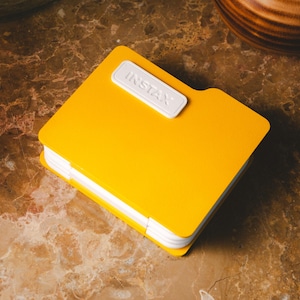 Op de afbeelding: Een heldergele Instax fotoalbum met een witte sluiting met het woord "INSTAX". Het vierkante album heeft witgerande pagina's. Het ligt op een bruin marmeren oppervlak.
