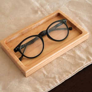 May include: Black-framed eyeglasses rest on a light-colored wooden tray. The tray is rectangular with raised edges and sits on a beige fabric surface. The glasses have a classic round shape.