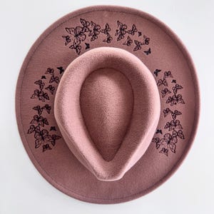 May include: A dusky pink fedora hat with a wide brim and a teardrop crown. The hat has a black butterfly design embroidered around the brim. The hat is made of a soft, felt-like material.