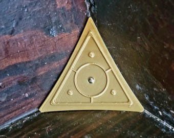 May include: A gold-colored triangular metal plaque with a circle and three smaller circles inside. The plaque is mounted on a wooden surface.