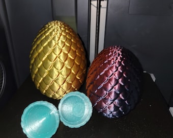May include: Three 3D printed dragon eggs, one gold, one purple, and one teal. The eggs are sitting on a black surface.